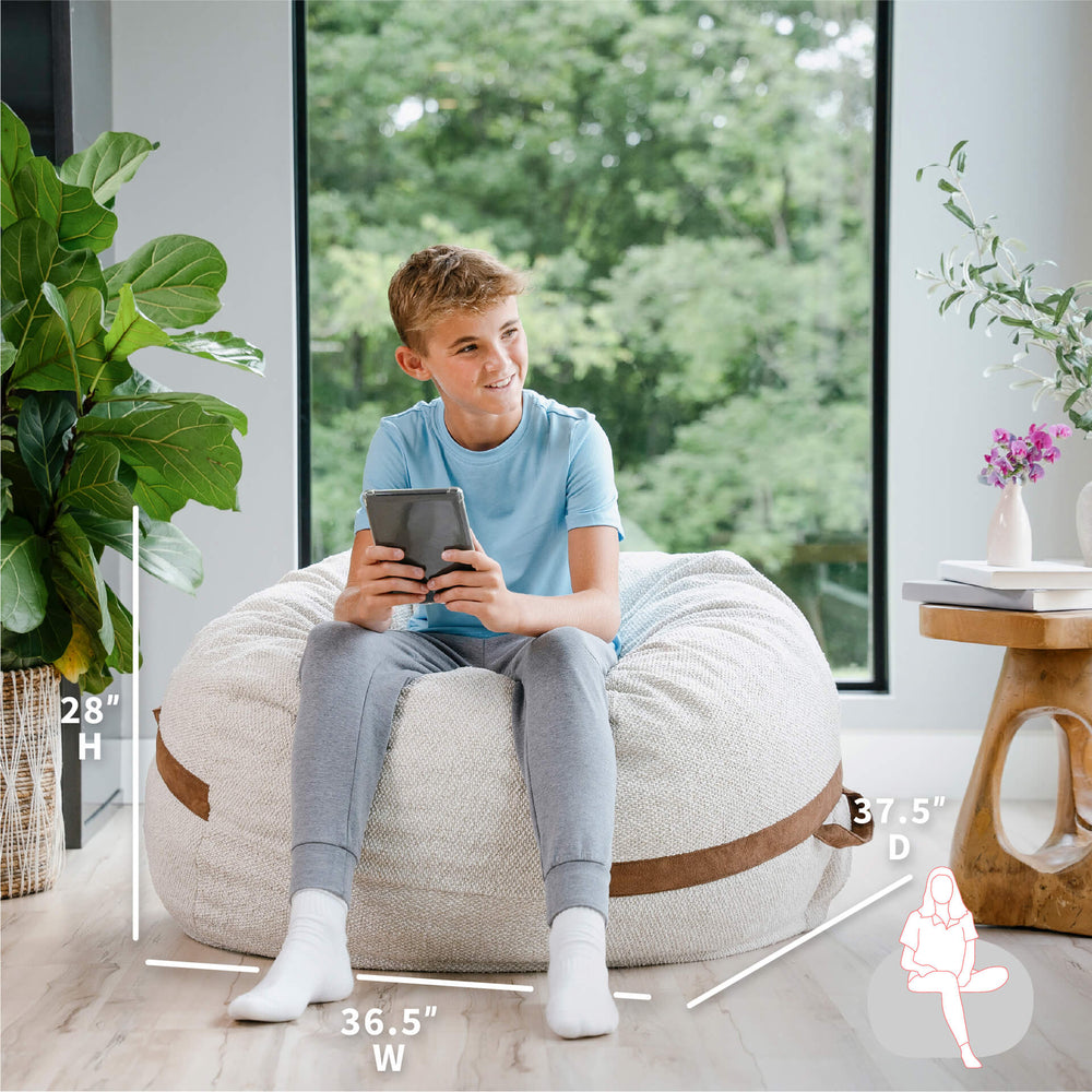 Young boy sitting on a bean bag chair holding a tablet, with measurements indicated around him. #color_sandstone-textured-boucle