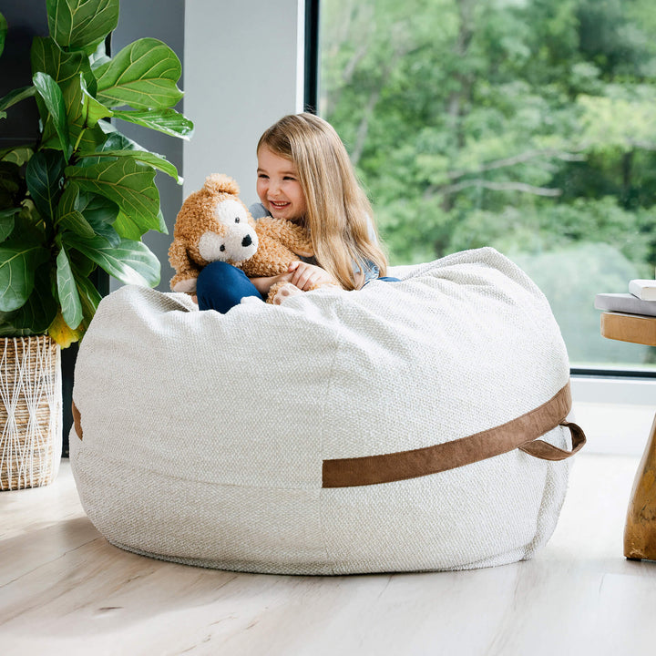 Child sitting in a bean bag chair holding a teddy bear in a bright room with large windows. #color_sandstone-textured-boucle