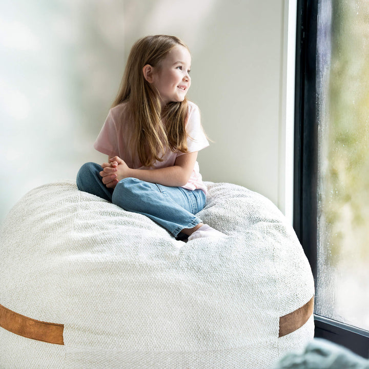 Young girl sitting on a large bean bag chair by a window #color_sandstone-textured-boucle
