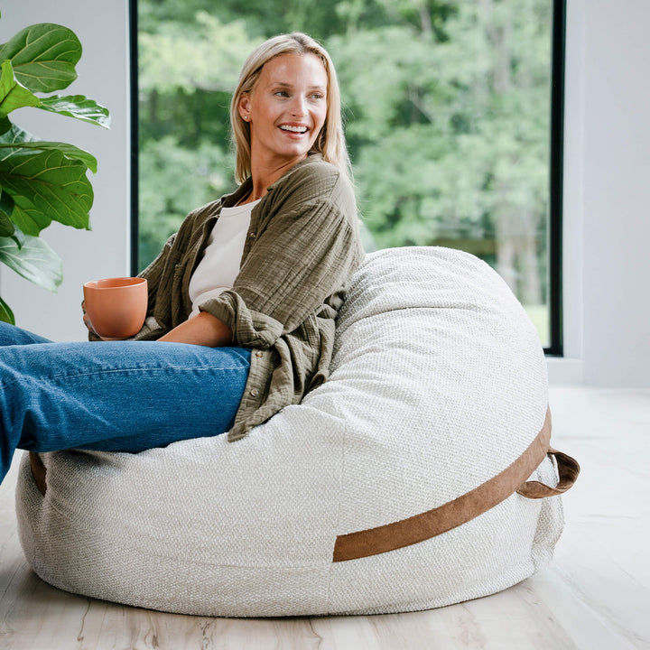 Woman sitting on a bean bag chair in a bright room with large windows and greenery. #color_sandstone-textured-boucle
