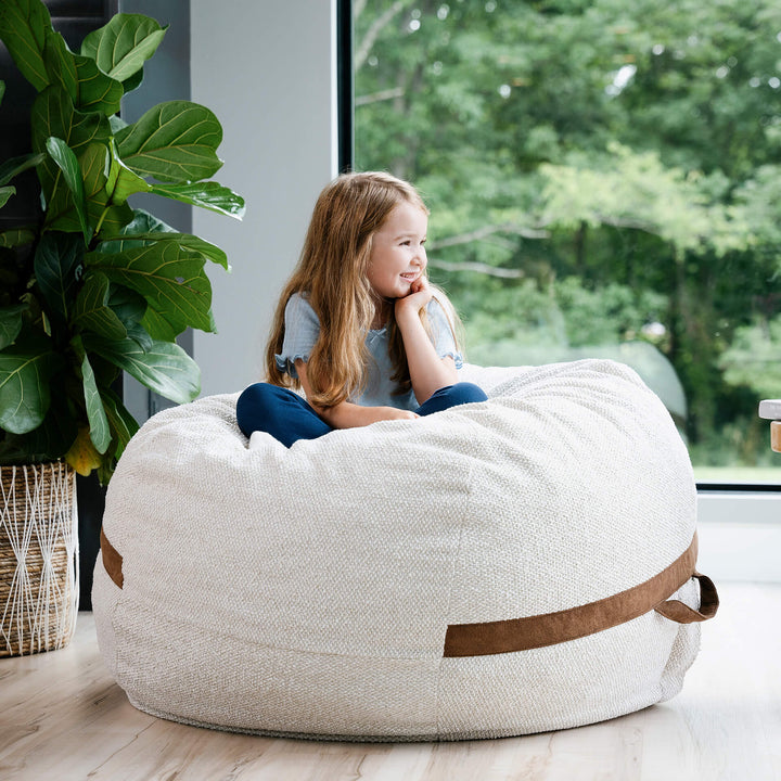 Child sitting on a bean bag chair in a room with a large window and plant. #color_sandstone-textured-boucle