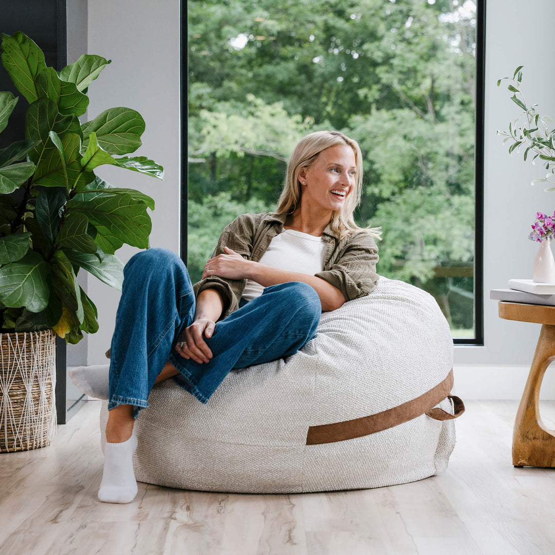 Woman sitting on a bean bag chair in a bright room with large windows and plants. #color_sandstone-textured-boucle