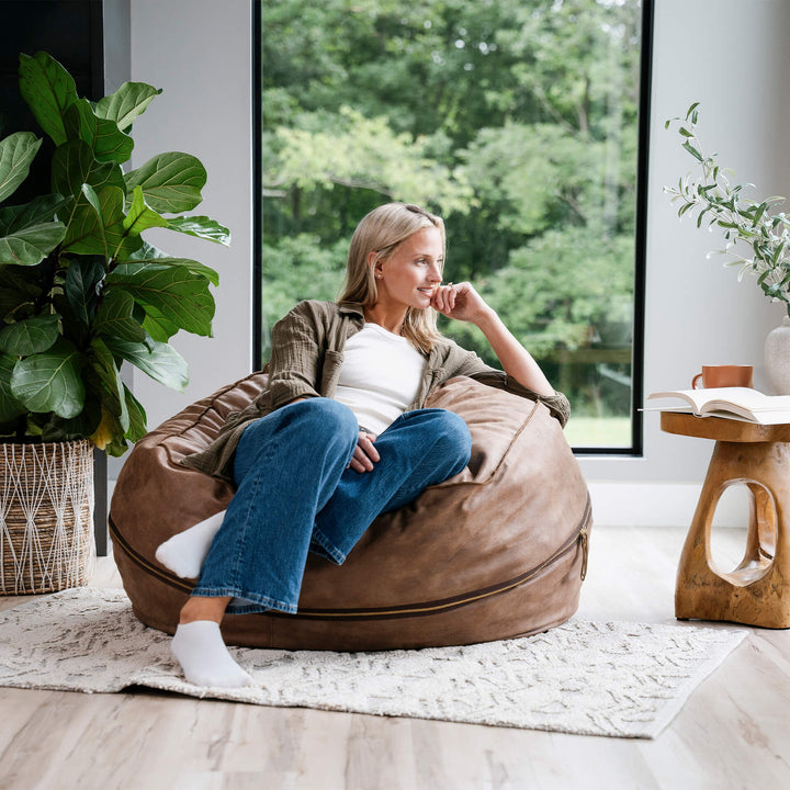 Woman sitting on a brown bean bag chair in a modern living room with large windows. #color_coffee-soft-vegan-leather