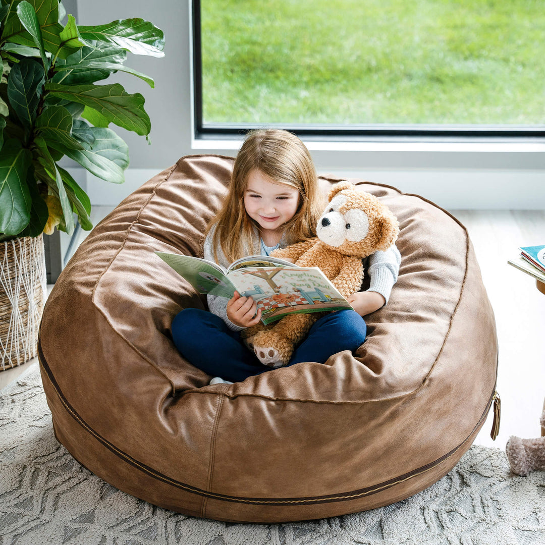 Child reading a book with a teddy bear on a large brown bean bag chair. #color_coffee-soft-vegan-leather