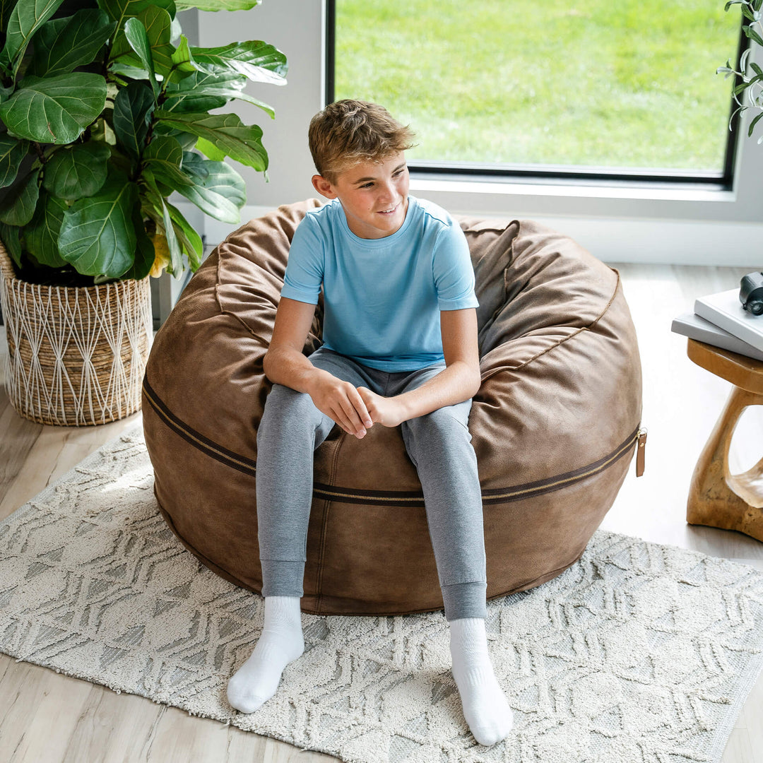 Child sitting on a brown bean bag chair in a room with a plant and window. #color_coffee-soft-vegan-leather