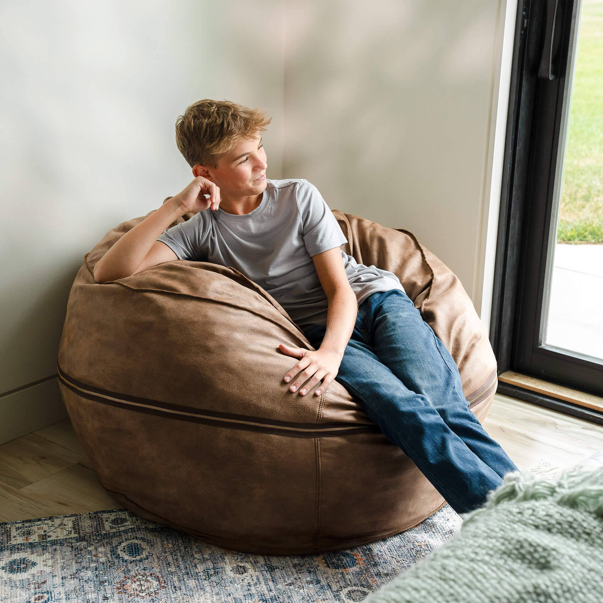 Person sitting on a brown bean bag chair in a room with a window. #color_coffee-soft-vegan-leather