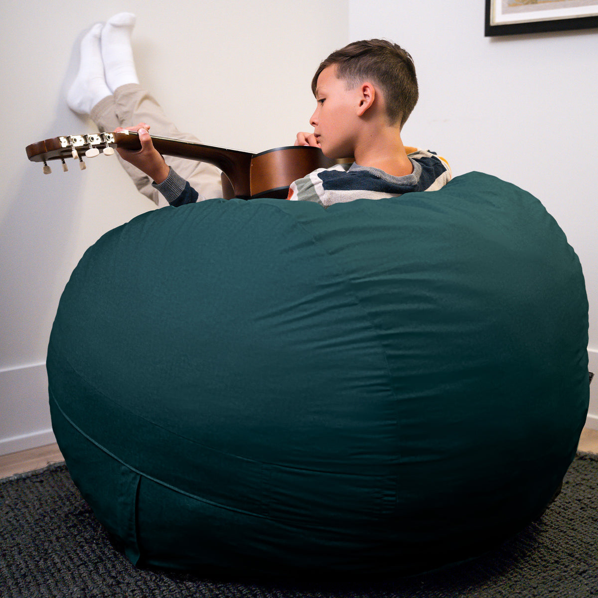 Person sitting on a large teal bean bag holding a guitar in a room with a white wall and framed picture. 