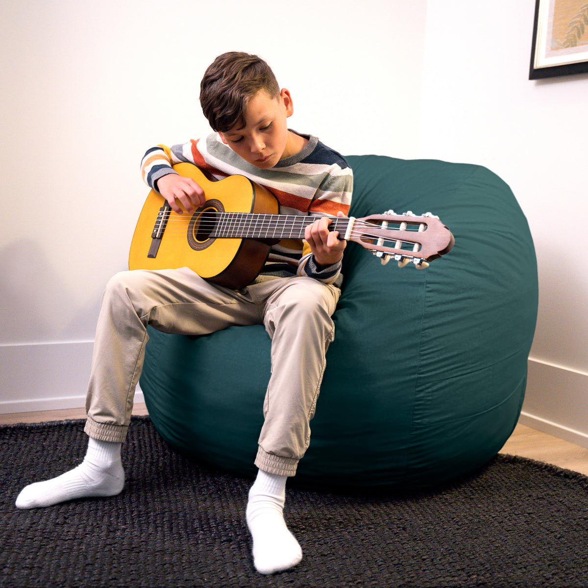 Child playing guitar on a large teal bean bag chair indoors. 