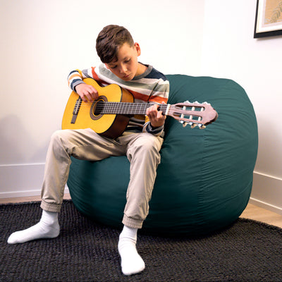 Child playing guitar on a large teal bean bag chair indoors. 