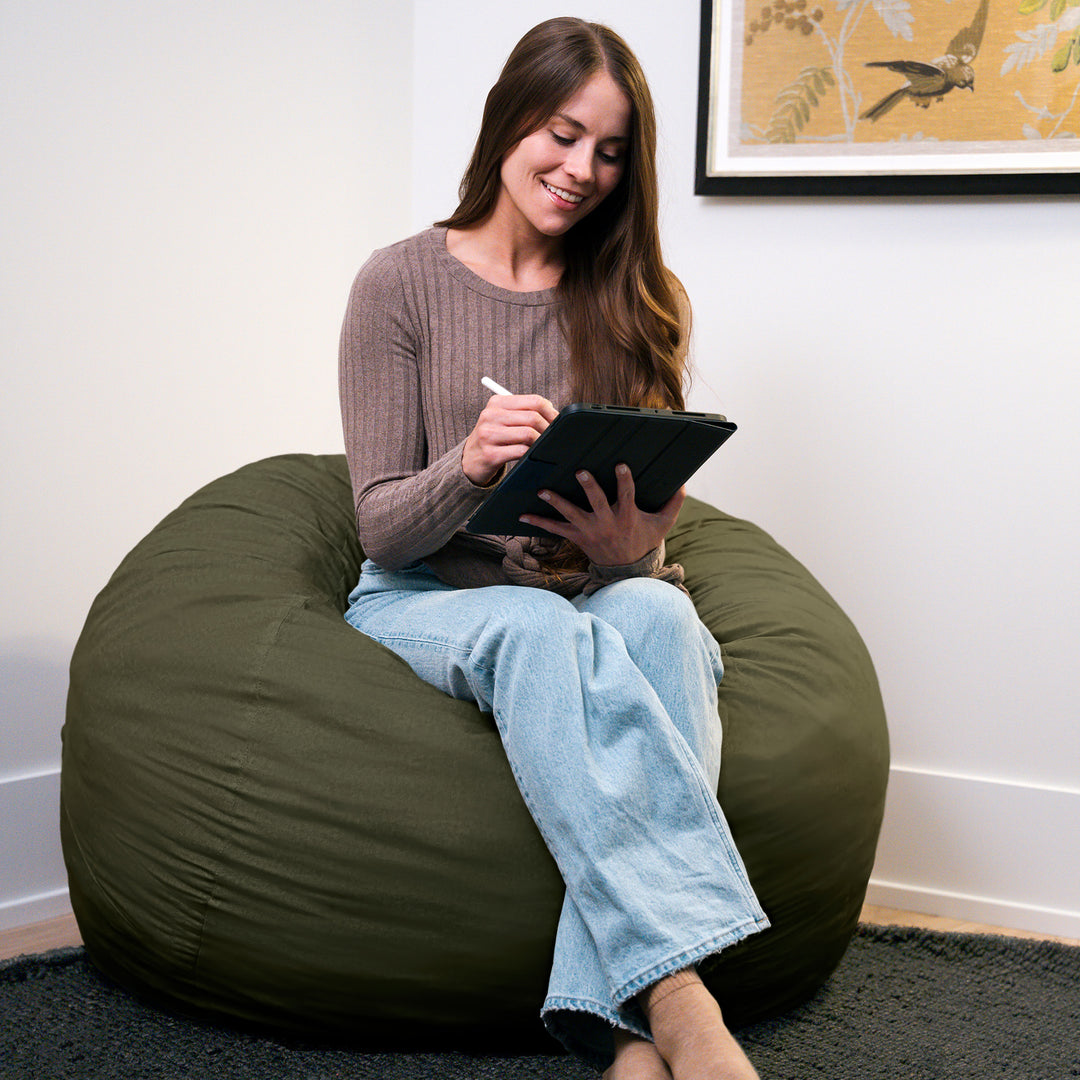 Woman sitting on a large green bean bag chair using a tablet. #color_forest-green-velveteen