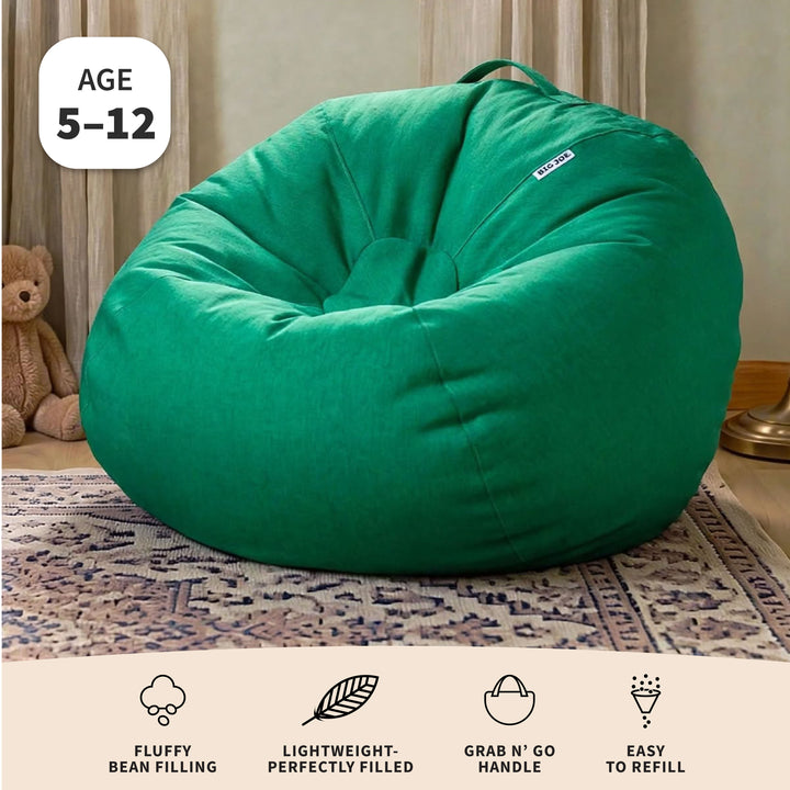 A large green bean bag chair shown in a styled indoor room setting with a rug, curtain, teddy bear, and lamp visible. The product is large and plush, with a small white brand tag on the upper right side. 