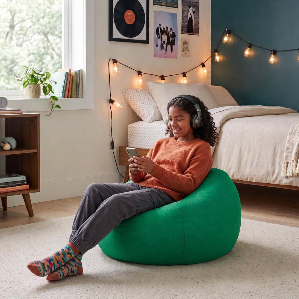 A woman wearing over-ear headphones sits smiling in a bright green bean bag chair in a cozy bedroom setting. She is wearing an orange sweater, gray pants, and colorful socks, holding a smartphone.  #color_vibrant-green-everyday-play