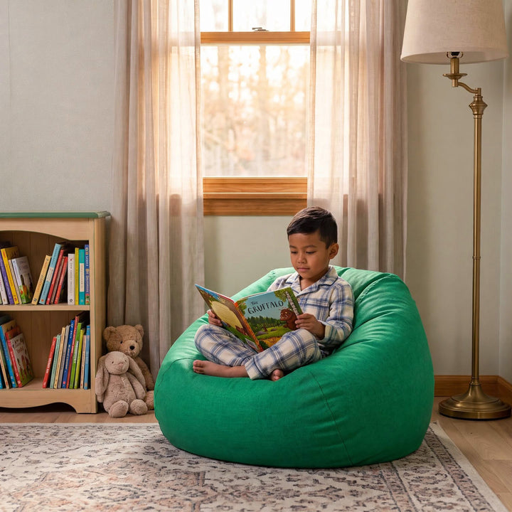 A child in plaid pajamas sits barefoot on a large green bean bag chair in a cozy room, reading a book. A bookshelf, window with sheer curtains, floor lamp, and patterned rug are visible. 