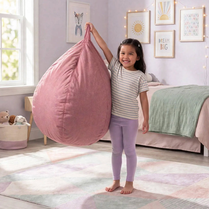 A young girl stands barefoot in a pastel bedroom, holding up a large dusty pink bean bag chair. The room features a bed with a green blanket, framed wall art, and a geometric pastel rug. 