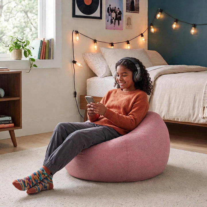 A woman sits smiling in a pink fabric bean bag chair in a cozy bedroom setting, wearing an orange sweater, gray pants, and colorful socks while looking at her phone. 