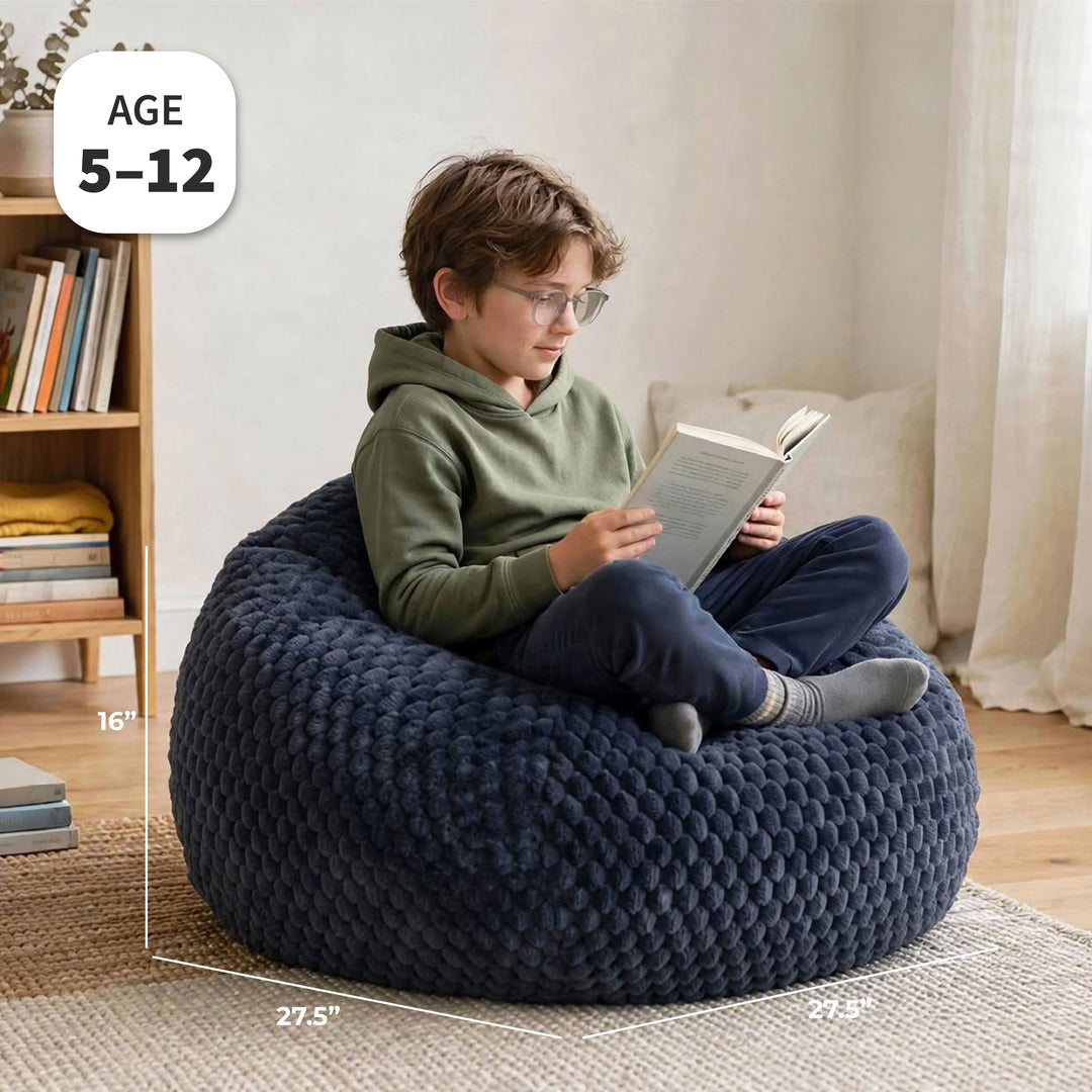 A child sitting on a large, chunky navy blue bean bag chair in a cozy room, reading a book. White dimension lines and text overlays show the chair&