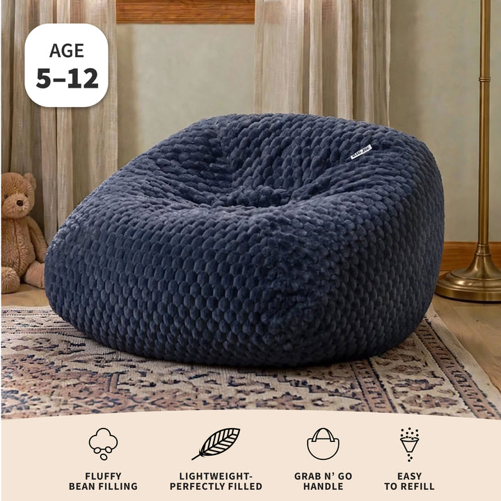 A large navy blue bean bag chair with a textured, honeycomb-like plush surface, shown in a styled room setting on a patterned rug. The image includes a white badge in the top left reading &