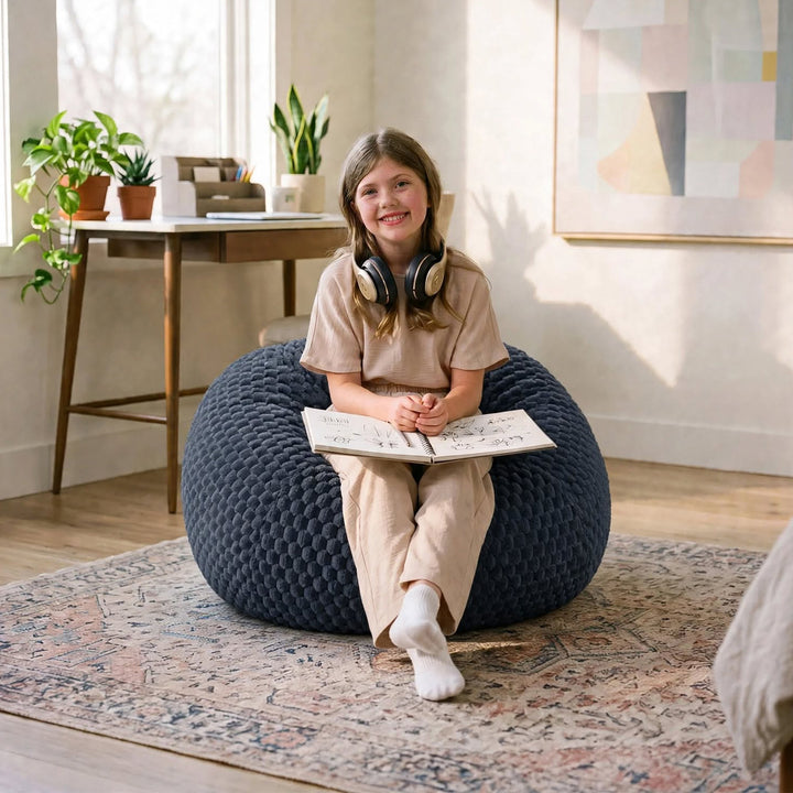 A young girl sits smiling on a large dark blue textured bean bag chair in a bright, modern room. She is wearing beige pajamas and has headphones around her neck while holding an open book. 