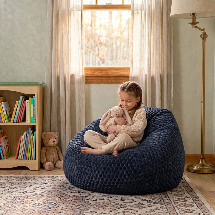 A young girl sits cross-legged on a large, textured navy blue bean bag chair in a cozy, sunlit children&