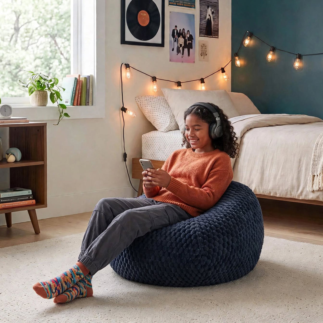 A woman sits smiling on a large navy blue chunky-knit bean bag chair in a cozy, modern bedroom. She is wearing a rust-orange sweater, gray pants, and colorful knit socks, holding a smartphone. 