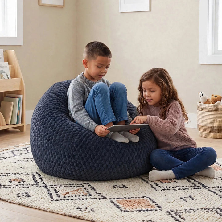 A boy and a girl sit on a large, chunky navy blue bean bag chair in a cozy, neutral-toned room. The boy wears a gray long-sleeve shirt and blue jeans, while the girl wears a dusty pink long-sleeve shirt and blue jeans. 