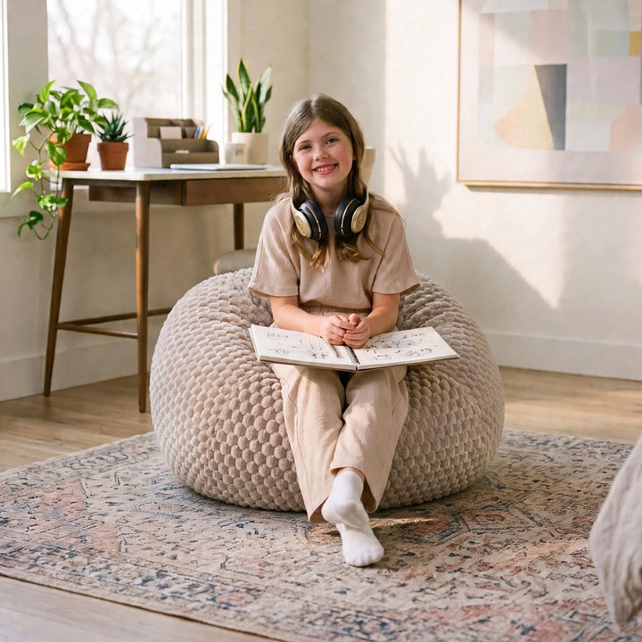 A young girl with headphones around her neck sits smiling on a large, textured beige bean bag chair in a bright, modern room. She is wearing a matching beige short-sleeve top and pants set, sitting on a patterned rug.  