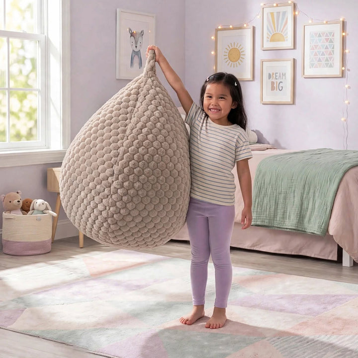 A young girl stands barefoot in a decorated bedroom, holding up a large, teardrop-shaped chunky knit bean bag chair. The room features pastel-colored walls, framed art, a bed with a green blanket, and a patterned rug.  