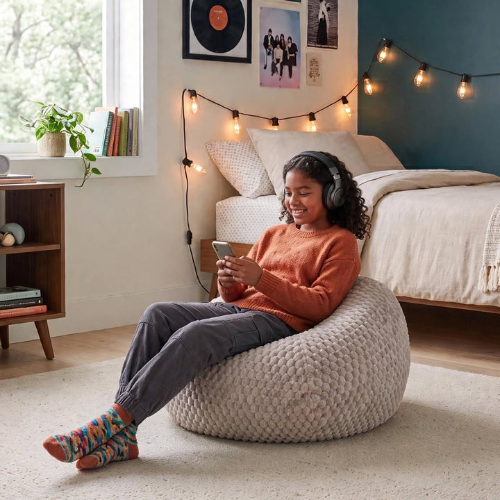 A woman wearing over-ear headphones sits smiling in a cozy bedroom while using her phone, lounging on a chunky knit beige bean bag chair. The room features a bed with cream bedding, a wooden shelf, and warm string lights.  