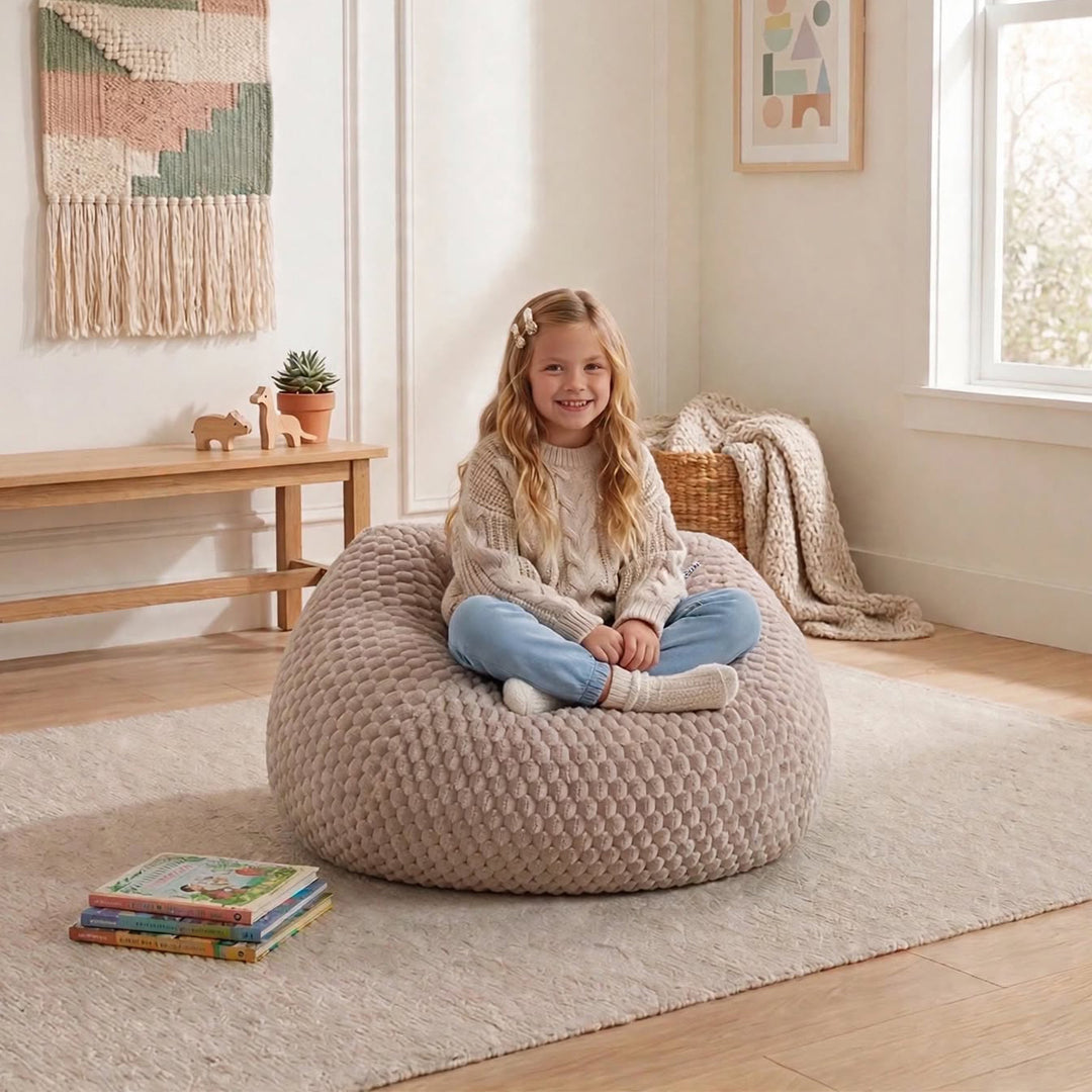 A young girl sits cross-legged on a large, textured beige bean bag chair in a bright, cozy room with light wood floors, a rug, and home decor.  
