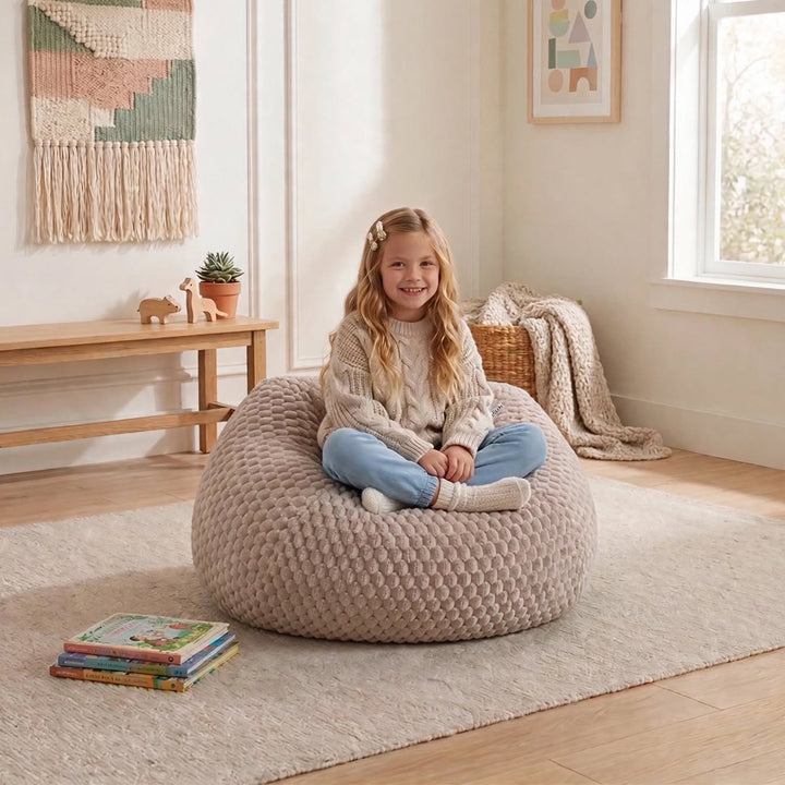 A young girl sits cross-legged on a large, textured beige bean bag chair in a bright, cozy room with light wood floors, a rug, and home decor.  