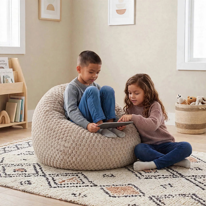 Two children sitting on a large beige textured bean bag chair in a bright, modern room, looking at a tablet. The room features a patterned rug, a wooden bookshelf, and a wicker basket with toys.  