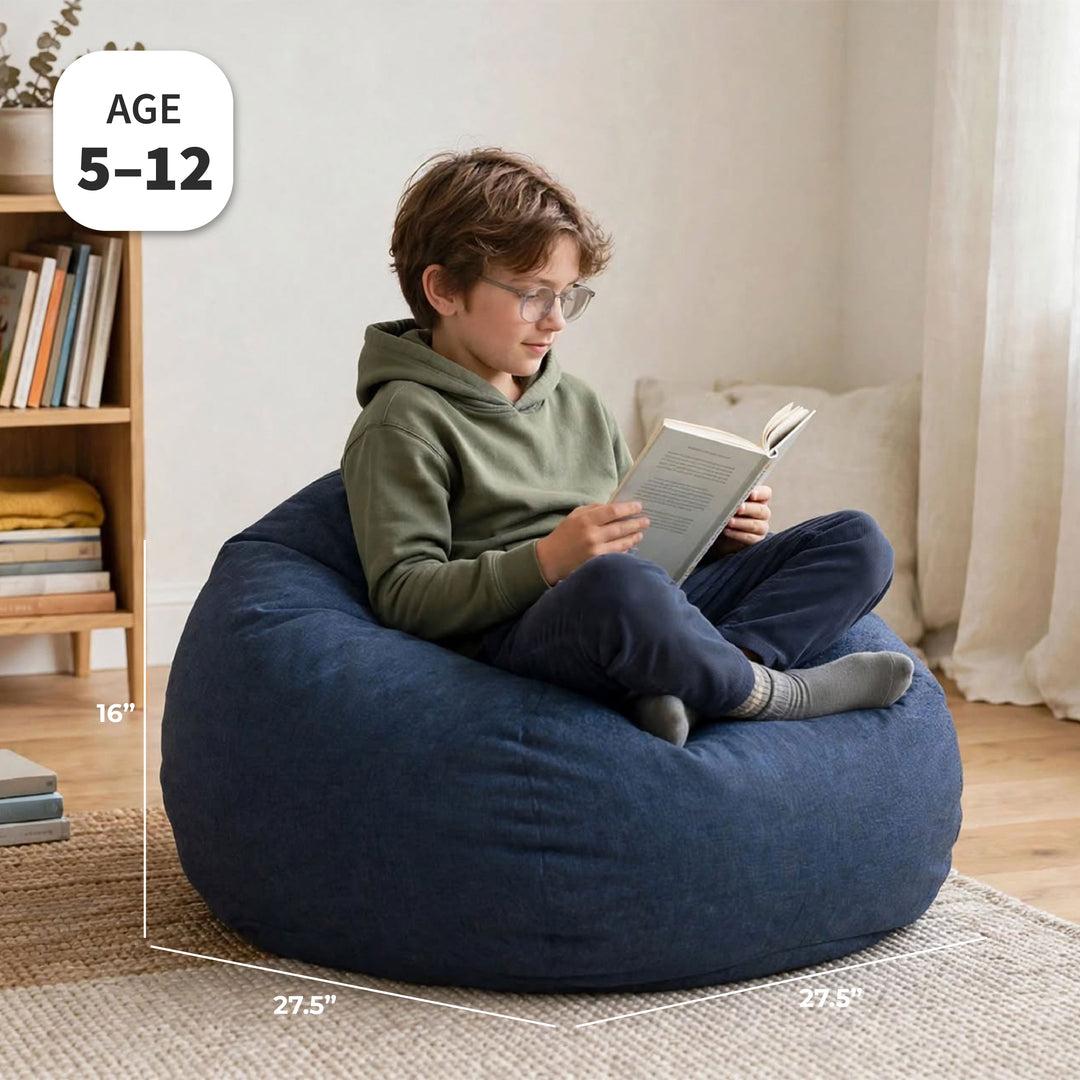 A child sitting cross-legged on a large navy blue bean bag chair in a bright room, reading a book. White dimension lines and text overlays show the chair&