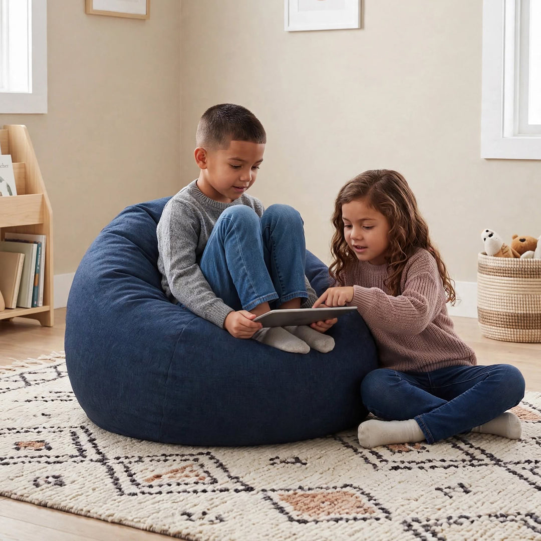Two children sitting on a large navy blue bean bag chair in a cozy, sunlit room. The boy wears a gray long-sleeve shirt and blue jeans, while the girl wears a dusty pink knit sweater, blue jeans, and white socks. 