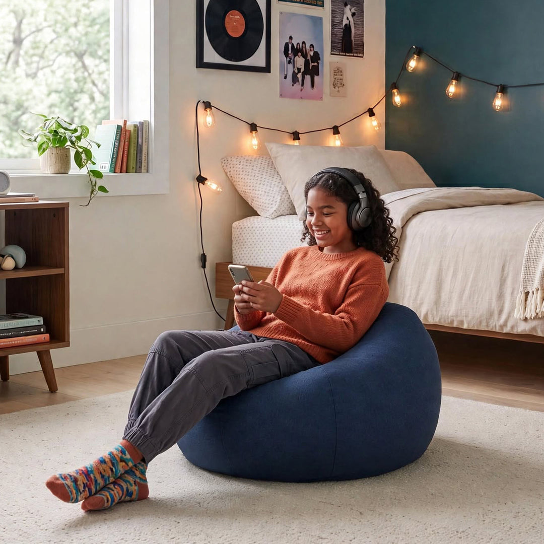 A woman wearing headphones sits relaxed in a large navy blue bean bag chair in a cozy, modern bedroom setting. She is wearing an orange knit sweater, gray pants, and colorful socks, holding a phone and looking down at it. 