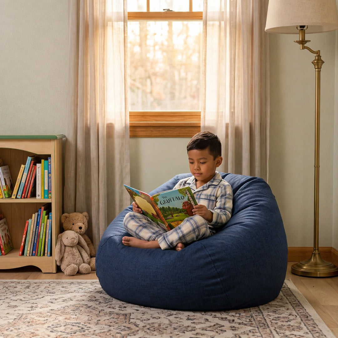 A young boy in a plaid pajama set sits barefoot in a large navy blue bean bag chair, reading a book in a cozy, sunlit child&