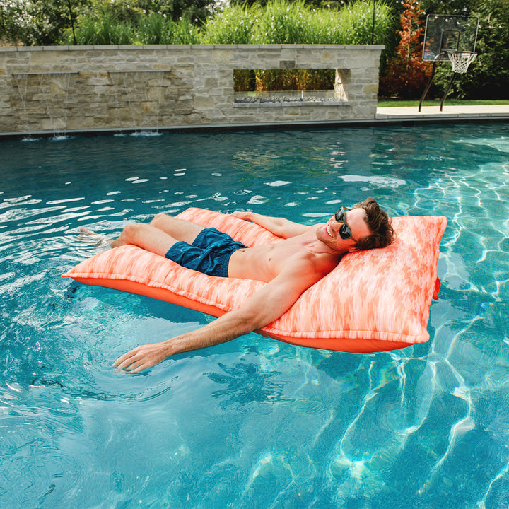 Man lying on a pink pool float in a swimming pool with a stone wall and greenery in the background. #color_peach-glow