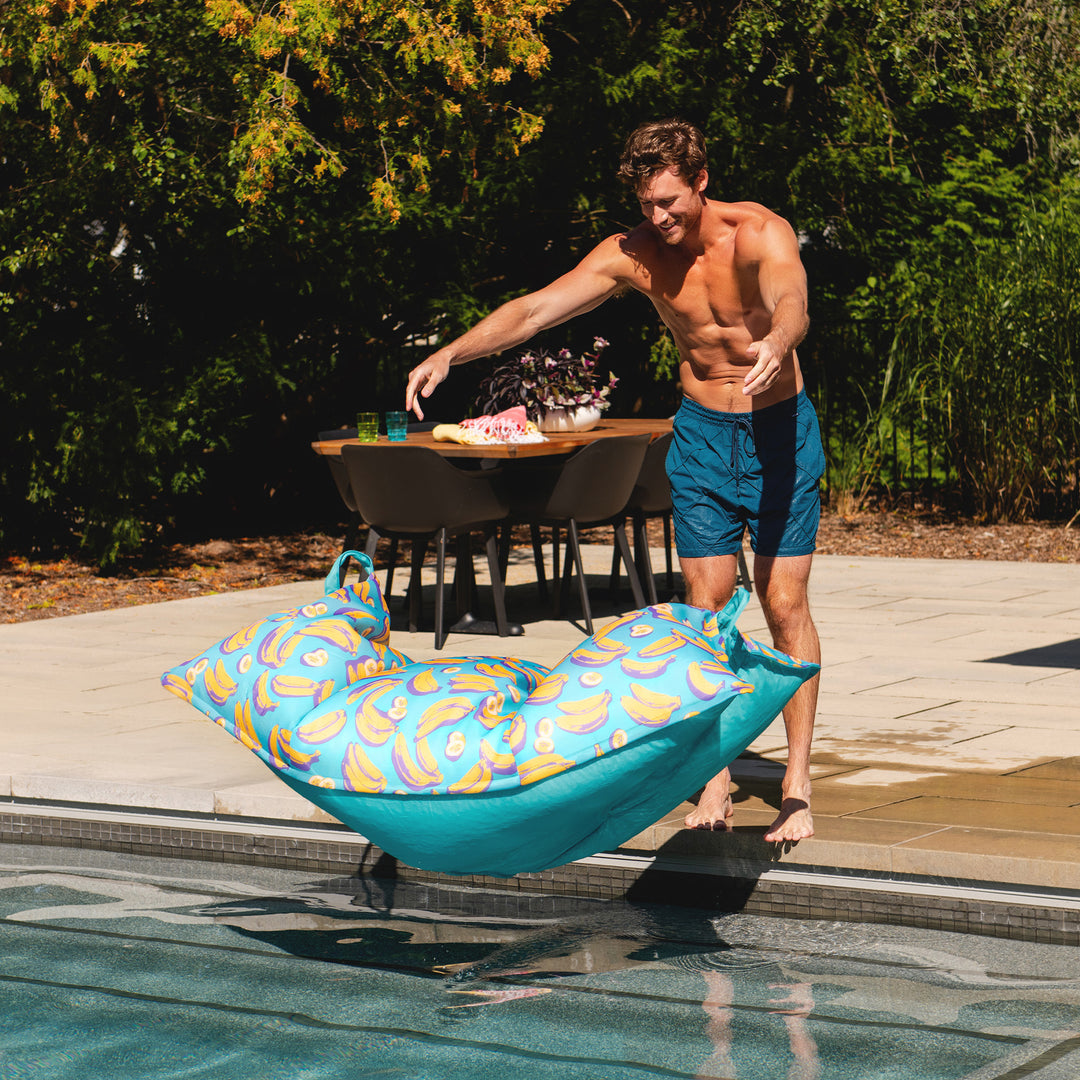Man holding a blue pool toy by a poolside with a dining area in the background #color_banana-pop