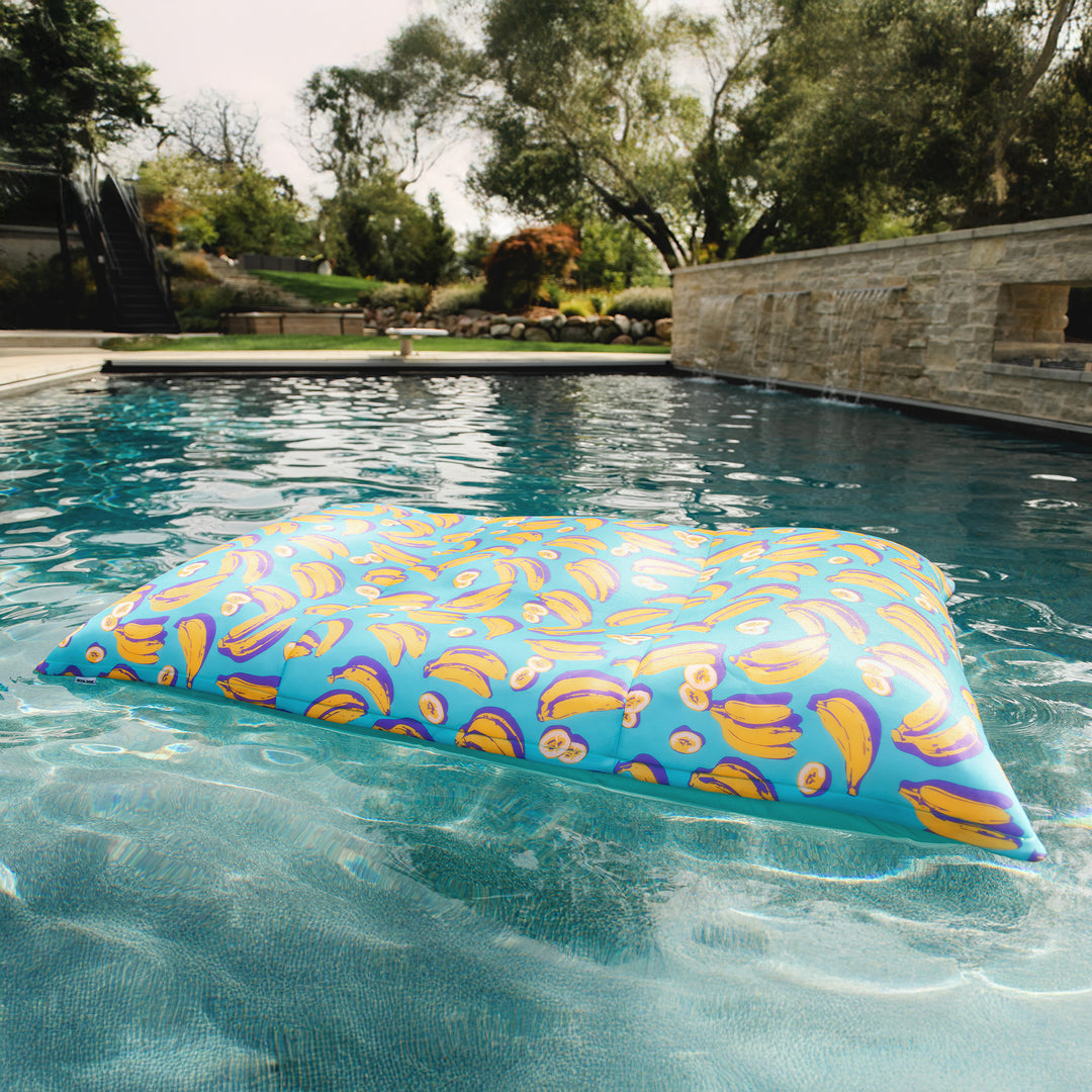 Colorful pool pillow floating in a pool with trees and a stone wall in the background. #color_banana-pop