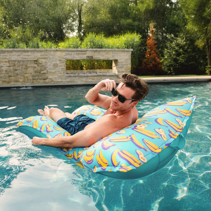 Man lounging on a banana-patterned pool float in a pool surrounded by greenery. #color_banana-pop