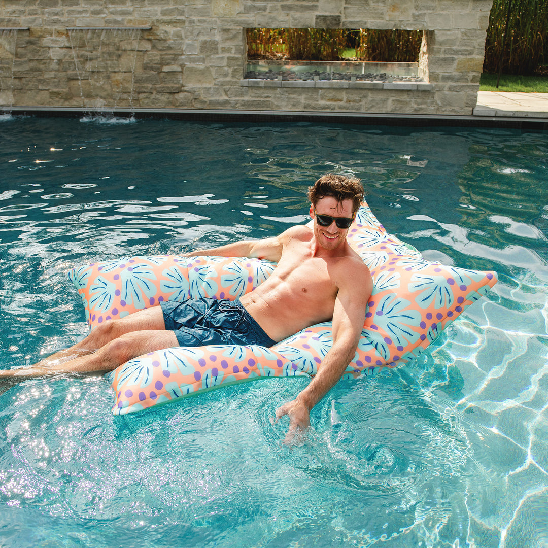 Man lounging on a floral raft in a pool. #color_sweet-shells-peach