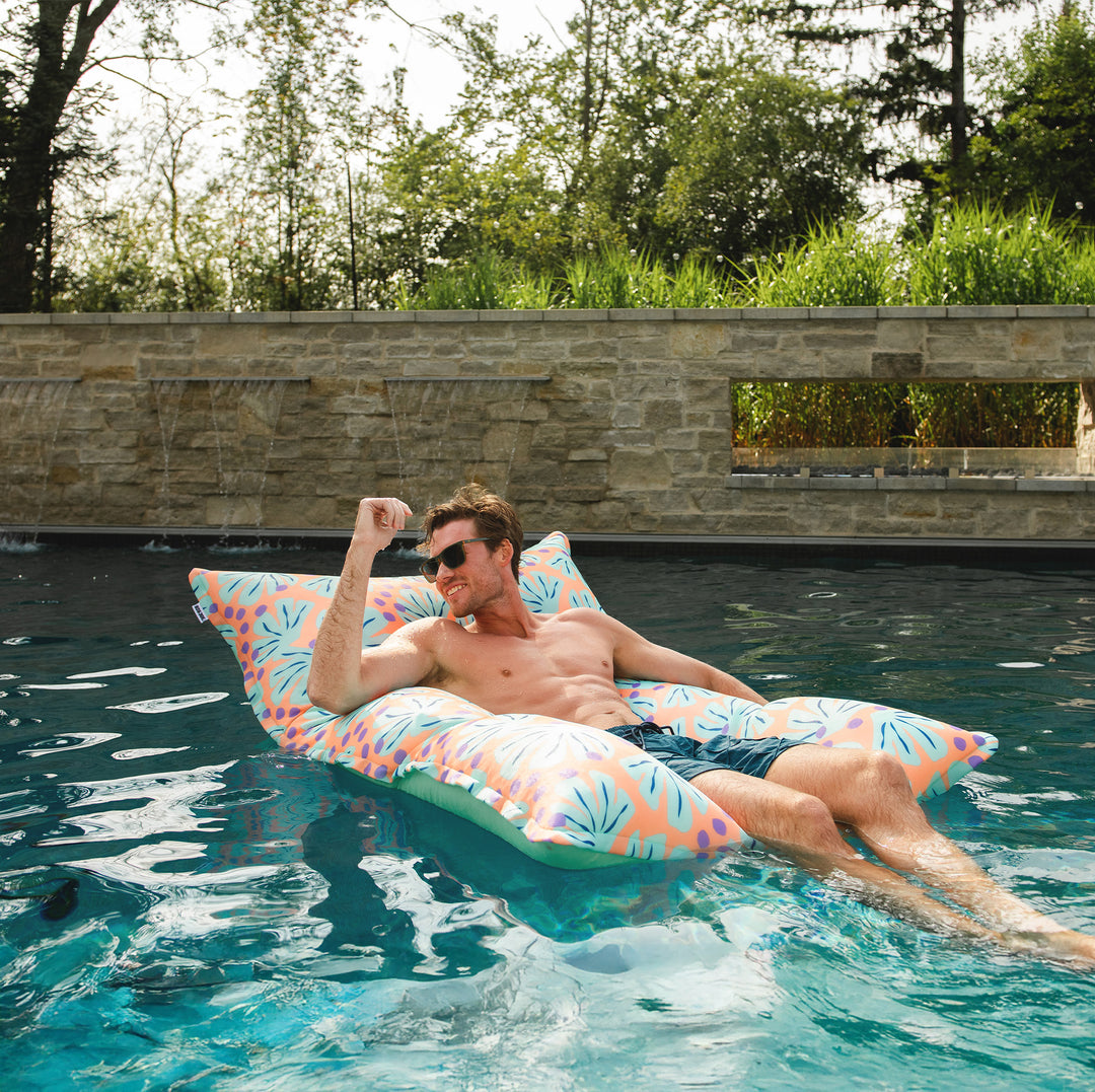 Man lounging on a colorful pool float in a pool with a stone wall and greenery in the background. #color_sweet-shells-peach