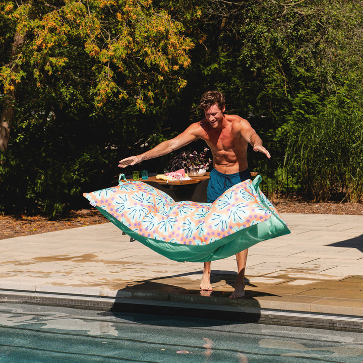 Man holding a colorful pool float by a poolside with a table in the background #color_sweet-shells-peach