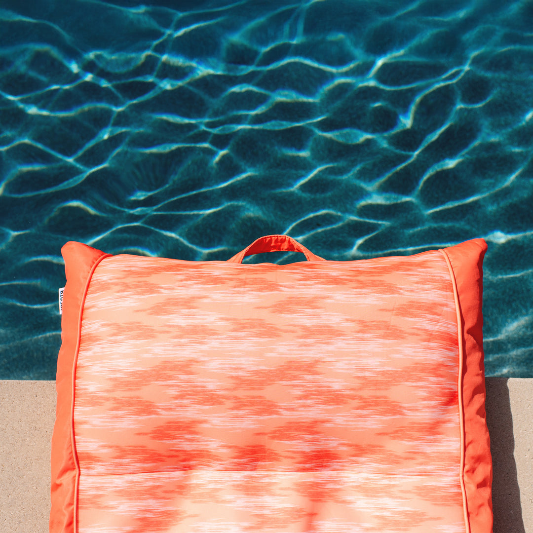 Orange and white patterned pool float on a sandy beach with clear blue water in the background #color_peach-glow