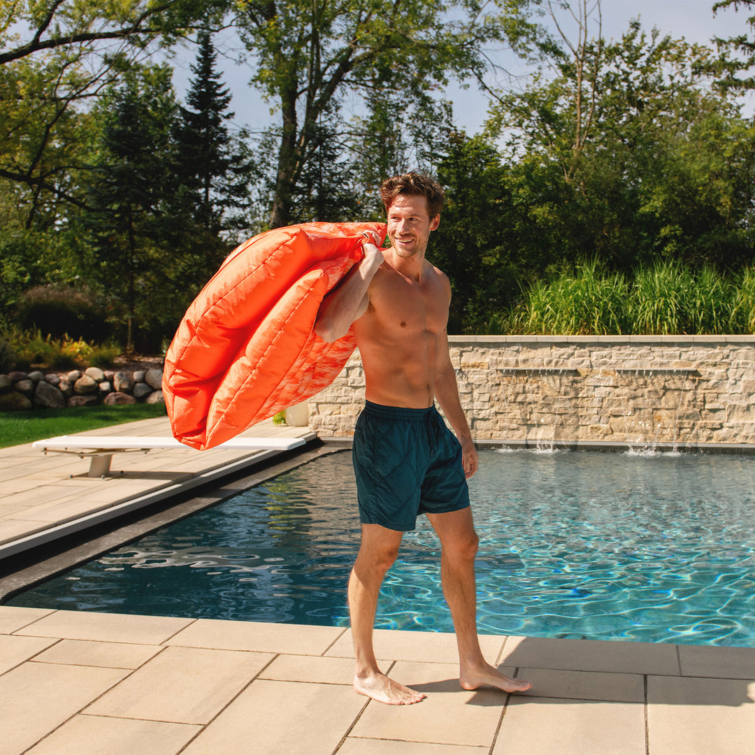 Man holding an orange pool lounger by a pool with trees in the background #color_peach-glow