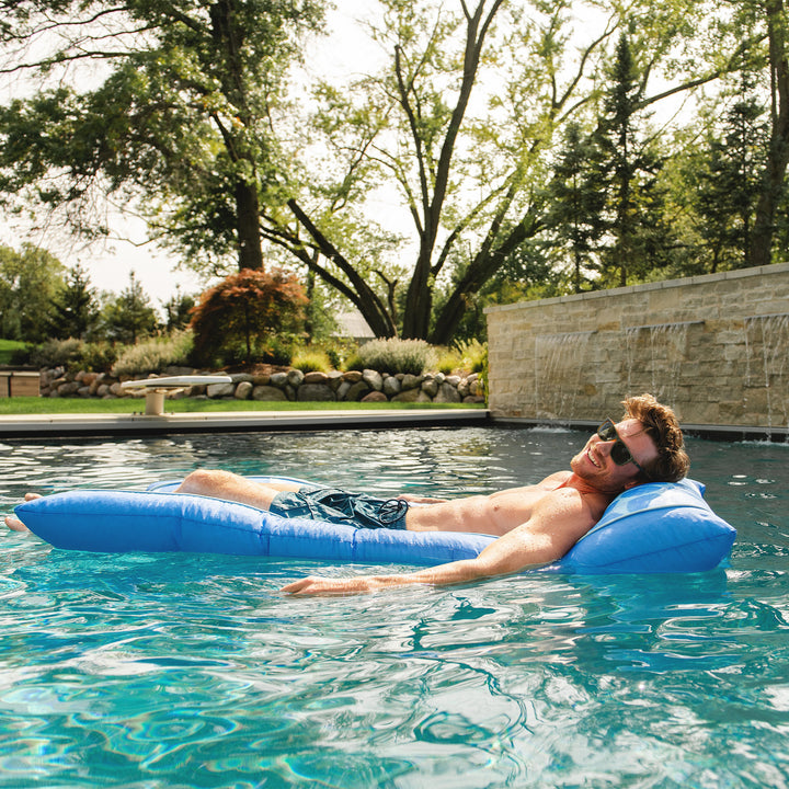 Man lying on a blue pool lounger in a pool with trees and stone wall in the background #color_coral-bloom