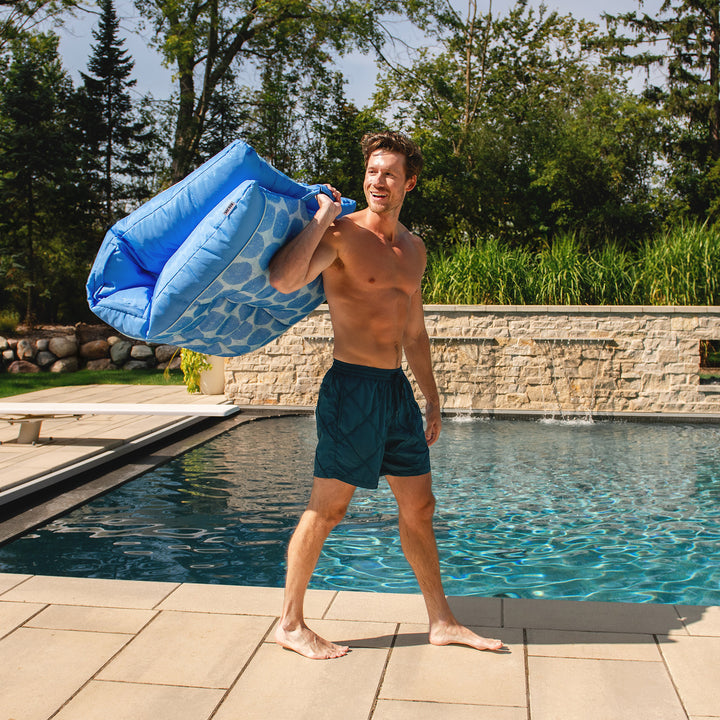 Man carrying a blue pool toy by a pool with trees in the background #color_coral-bloom