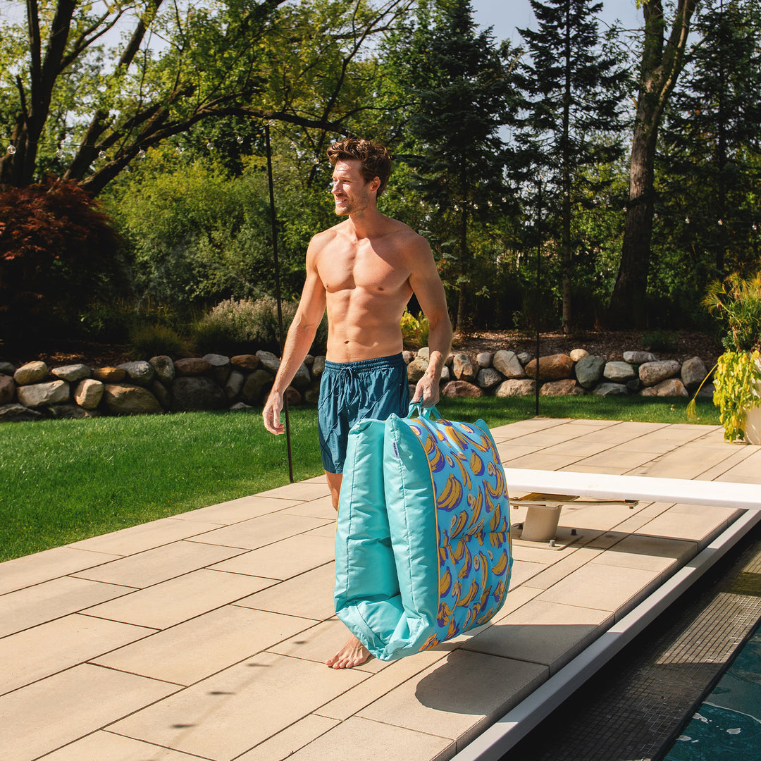 Man in swim shorts holding a blue and yellow pool float by a poolside with greenery in the background #color_banana-pop