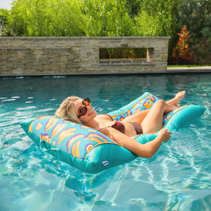 Woman relaxing on a colorful pool float in a pool with greenery in the background #color_banana-pop