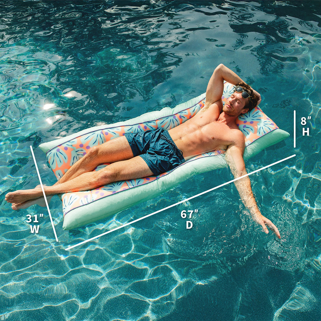 Man lying on a floral-patterned pool raft in clear blue water with measurements displayed. #color_sweet-shells-peach