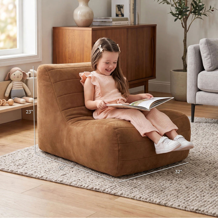 A young girl sits smiling in a brown upholstered armchair in a styled living room, reading a book. White dimension lines overlay the chair showing its 23 x 32 x 70 inch measurements. 